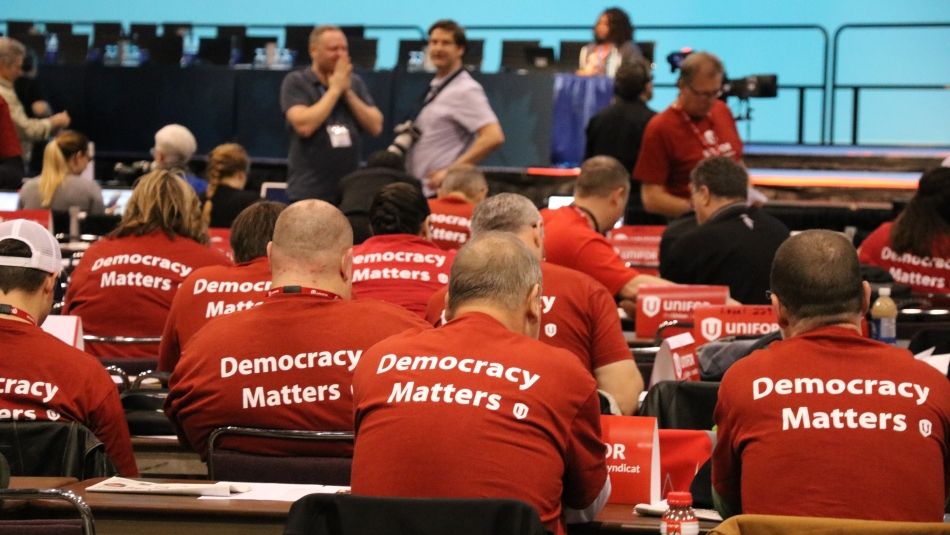 Seen from behind, a roomfull of Unifor members wearing matching "Democracy Matters" t-shirts.
